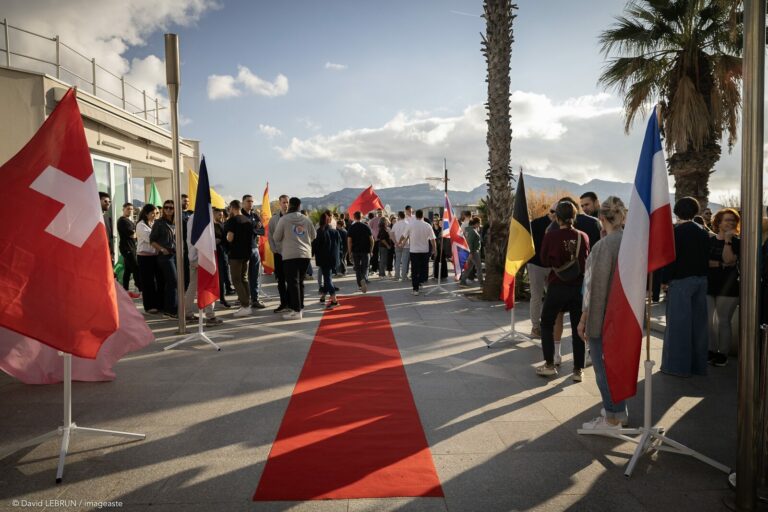 Arrivee gala convention GM 2025, tapis rouge et drapeaux nations, accueil collaborateurs en exterieur ensoleille